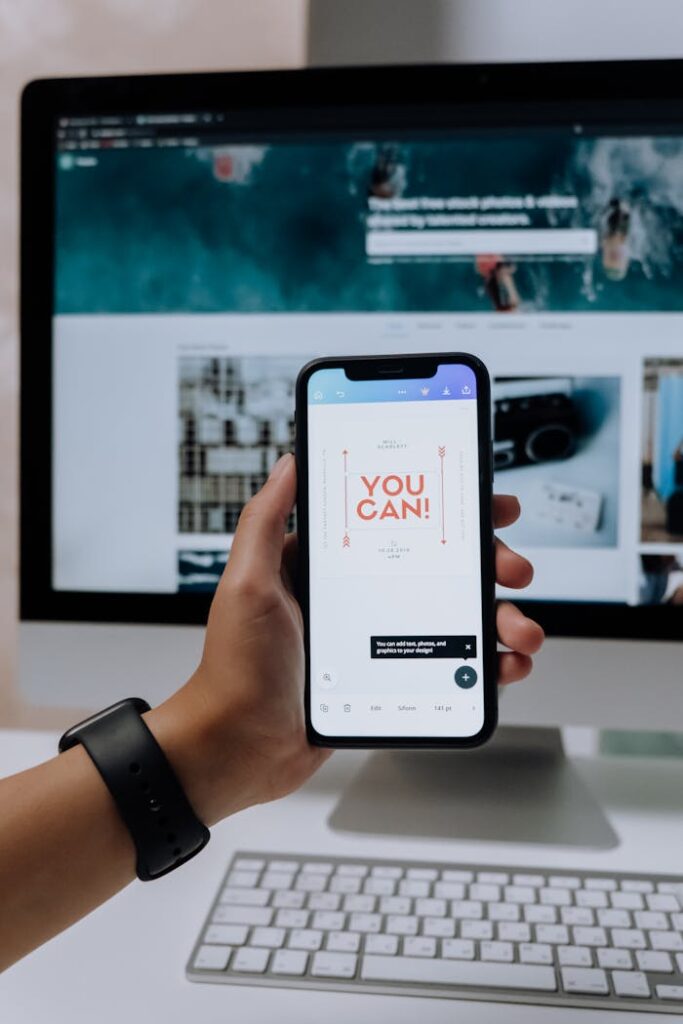 Hand holding smartphone with 'You Can!' message in front of desktop screen.